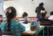 alunas crianças vistas de costas sentadas em carteiras em sala de aula e professora ao fundo