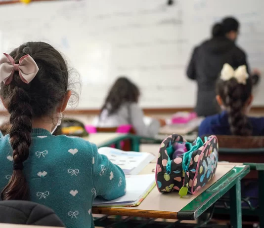 alunas crianças vistas de costas sentadas em carteiras em sala de aula e professora ao fundo