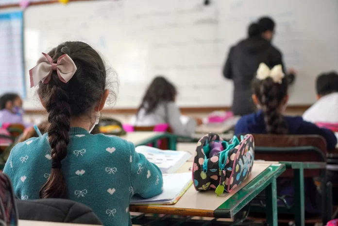 alunas crianças vistas de costas sentadas em carteiras em sala de aula e professora ao fundo